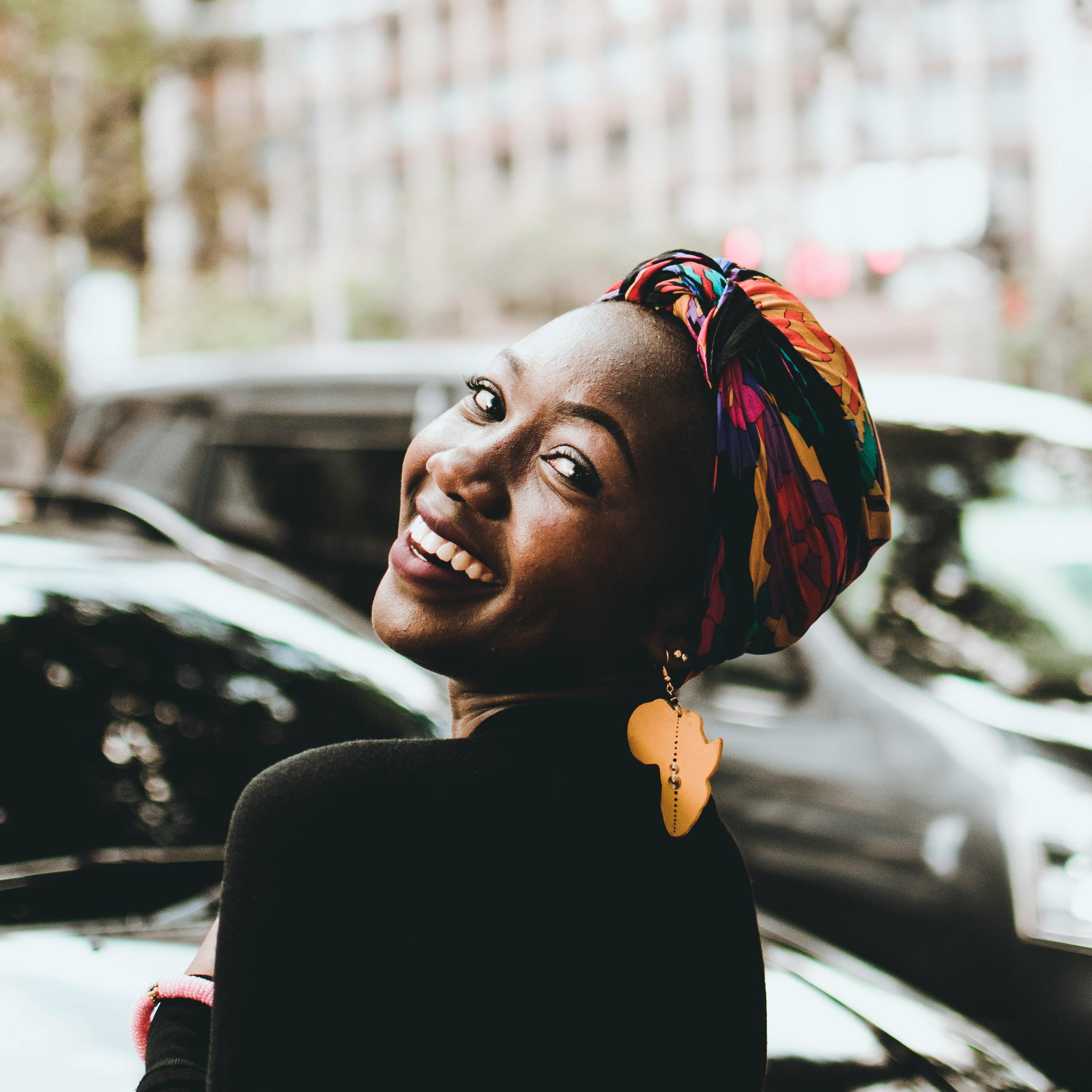 A joyful woman wearing a colorful headwrap and distinctive earrings, smiling in an urban setting.